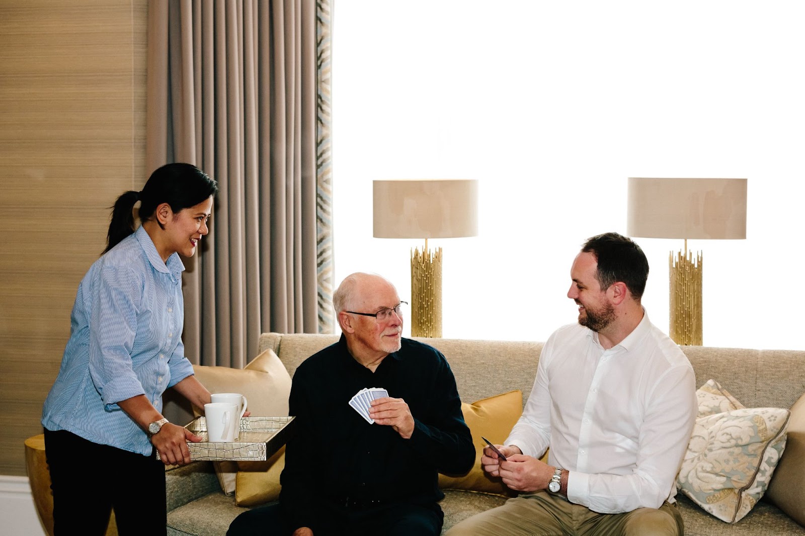 A caregiver serves tea to an elderly man with cards while a younger man sits nearby on a beige sofa in an elegant living room with warm lighting and curtains.