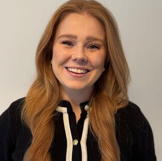 Smiling woman with long red hair wearing a black top with white trim, standing against a light background
